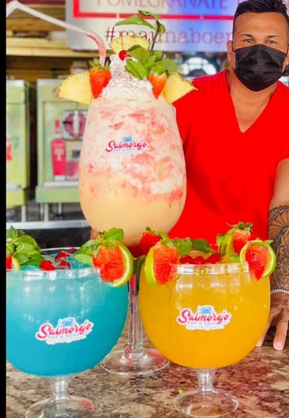 A bartender presenting three vibrant Salmorejo tropical cocktails, including a stacked pomegranate-flavored drink garnished with pineapple, strawberries, mint, and lime, alongside blue and orange versions on a bar counter, featuring the #mamaoboen hashtag and fresh fruit accents, perfect for Miami marina bar enthusiasts and cocktail lovers.